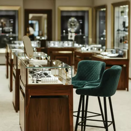 Interior view at Finnie's jewellery retail store in Aberdeen. In the foreground two green velvet bar chairs sit beside a counter with glass cabinet, ready for a consultation. Cabinets are dark wood with a glass display area on top. In the background the showroom recedes with further cabinets and display cases. Posters for luxury watches are just out of focus in the back of the shop.